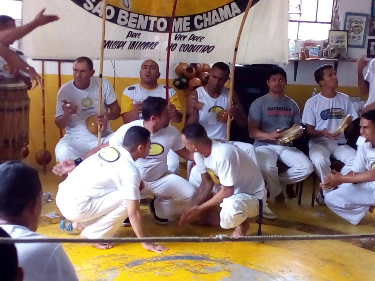 Dois capoeiristas agachados ao pé do berimbau, prontos para começar o jogo, com a bateria ao fundo