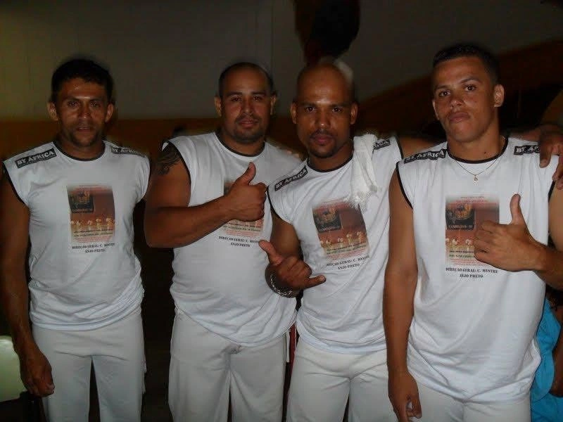 Quatro capoeiristas lado a lado com camisetas de um evento de capoeira