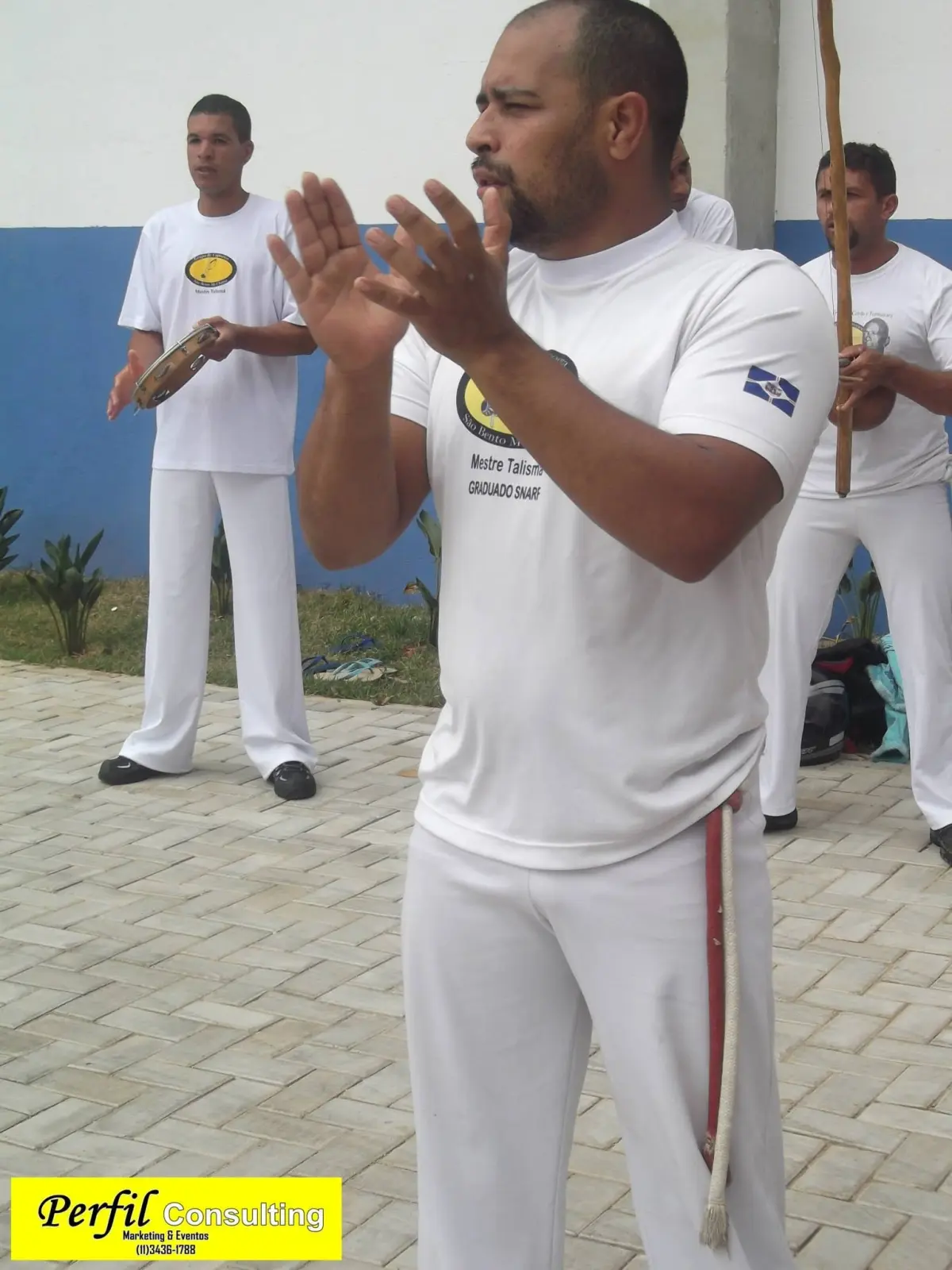 Capoeirista batendo palmas na roda ao ar livre, com pandeiro e berimbau ao fundo