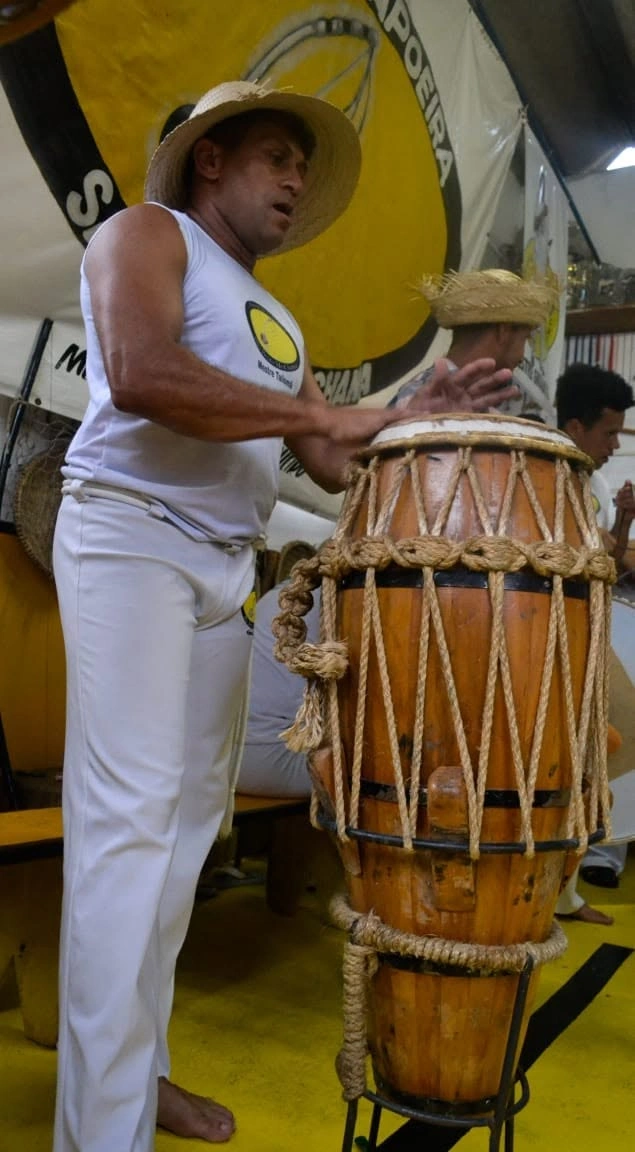 Capoeirista de chapéu de palha tocando atabaque em frente à faixa do grupo