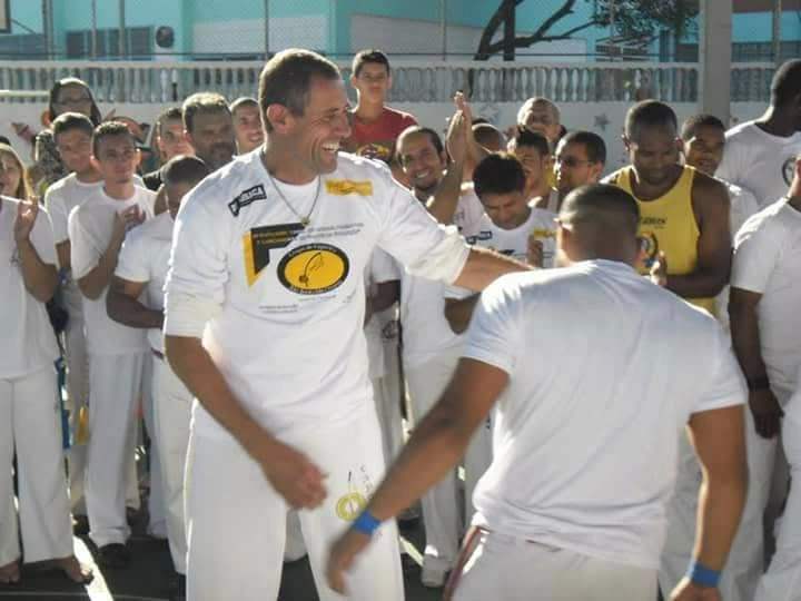 Capoeiristas sorrindo no centro de uma roda cheia, cercados por alunos de uniforme branco