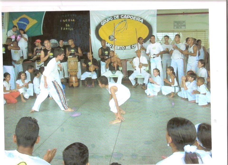 Roda de capoeira com crianças e adultos em volta, bateria ao fundo e a faixa do grupo