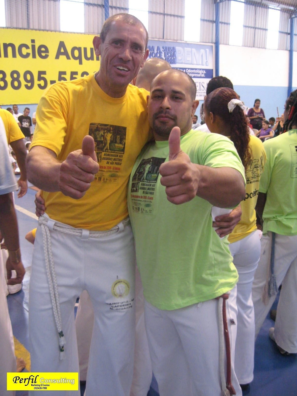 Dois capoeiristas abraçados fazendo sinal de positivo em evento de homenagem aos mestres de São Paulo